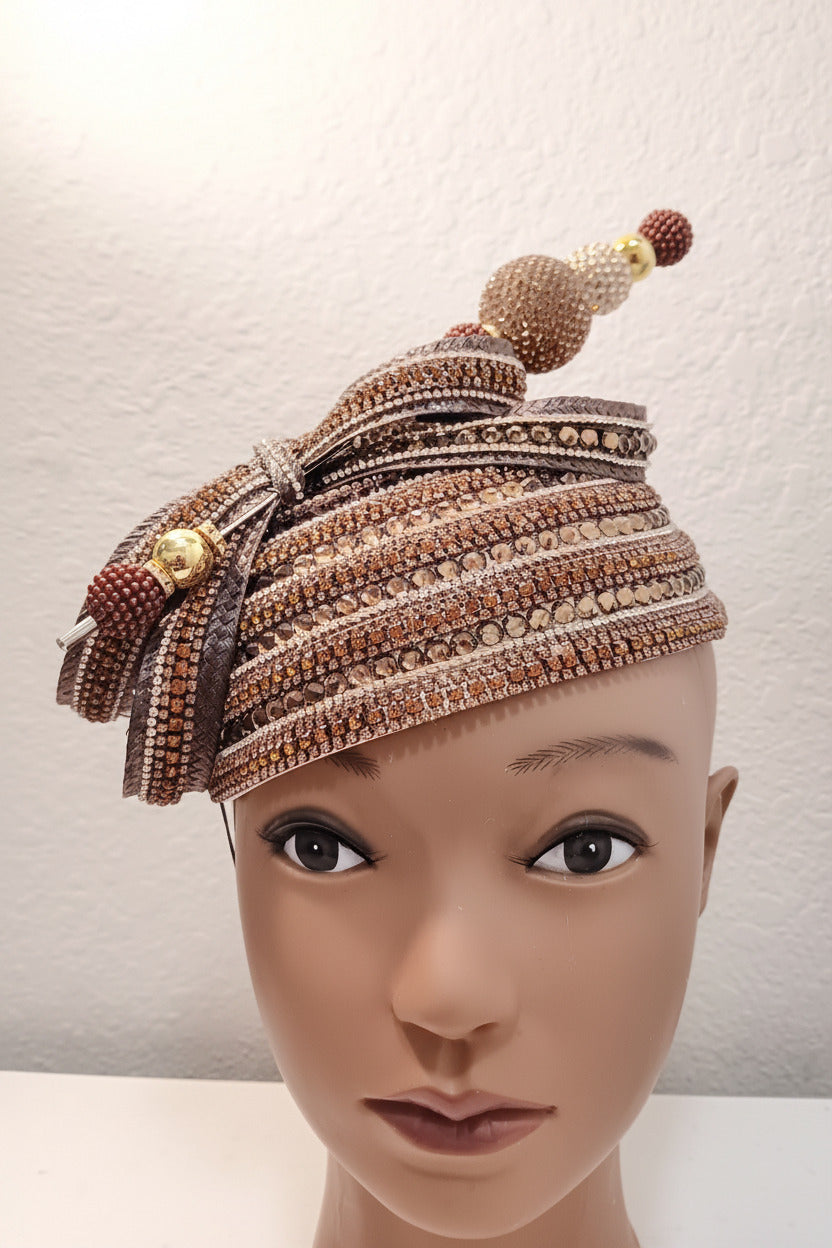 A highly detailed, ornate traditional African headwrap (agbada) embellished with intricate beadwork, sequins, and tassels, displayed on a mannequin head against a neutral textured wall.