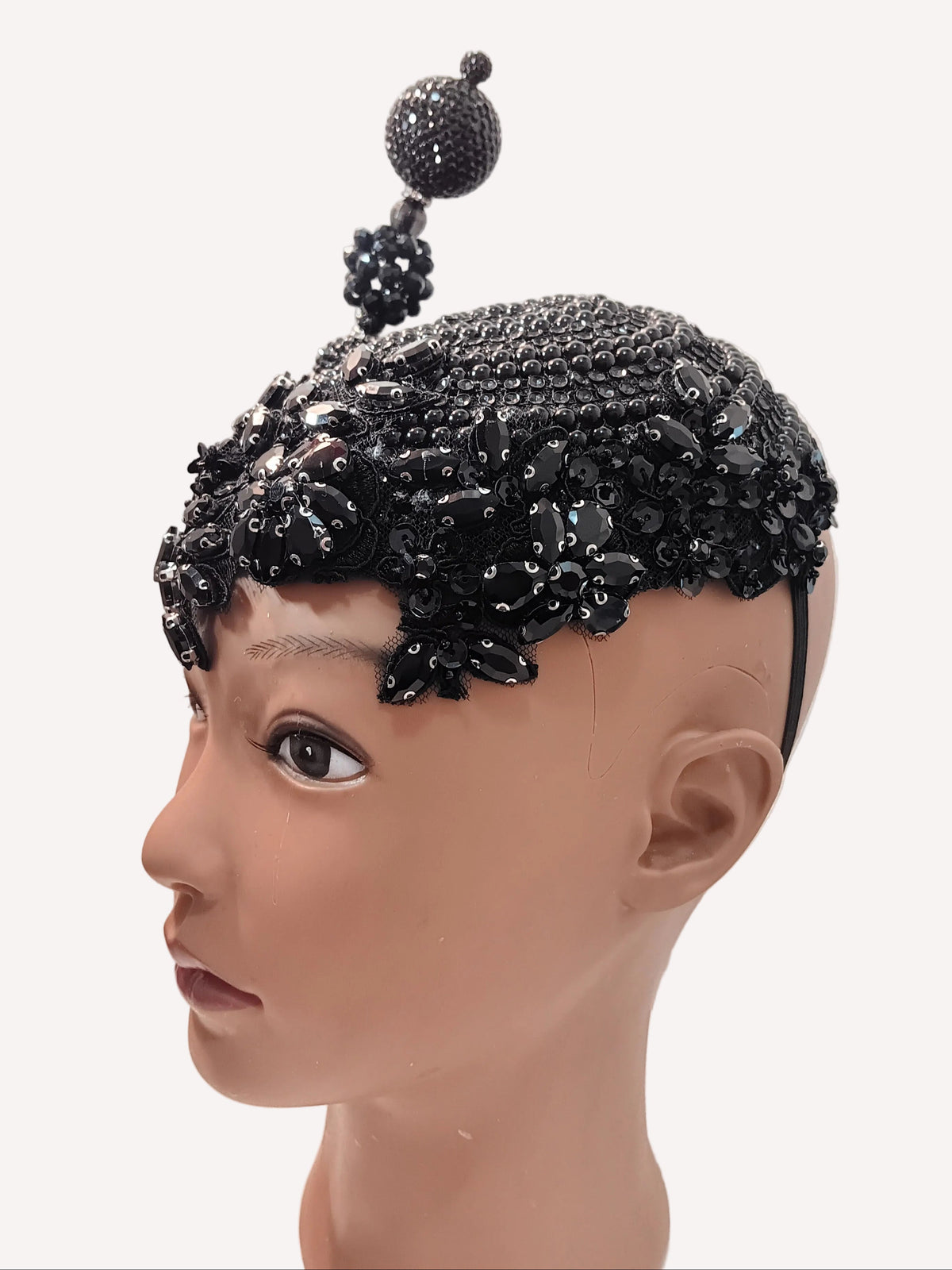 A black embellished headpiece with a tall central bead-topped spike, displayed on a mannequin head against a plain off-white wall.