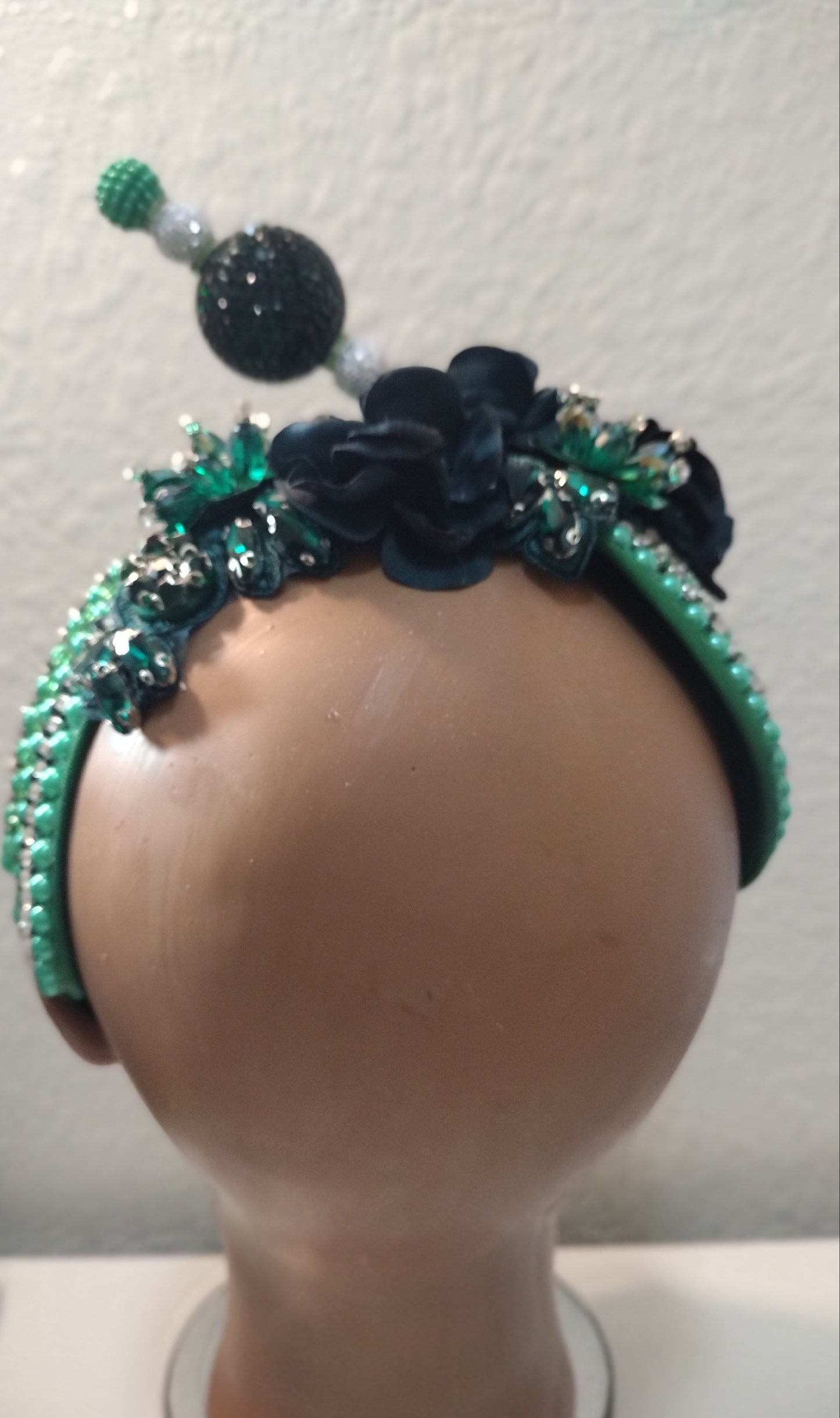 A decorative headband displayed on a mannequin head. The headband features dark navy blue fabric flowers, green beaded trim, and clear rhinestone embellishments.