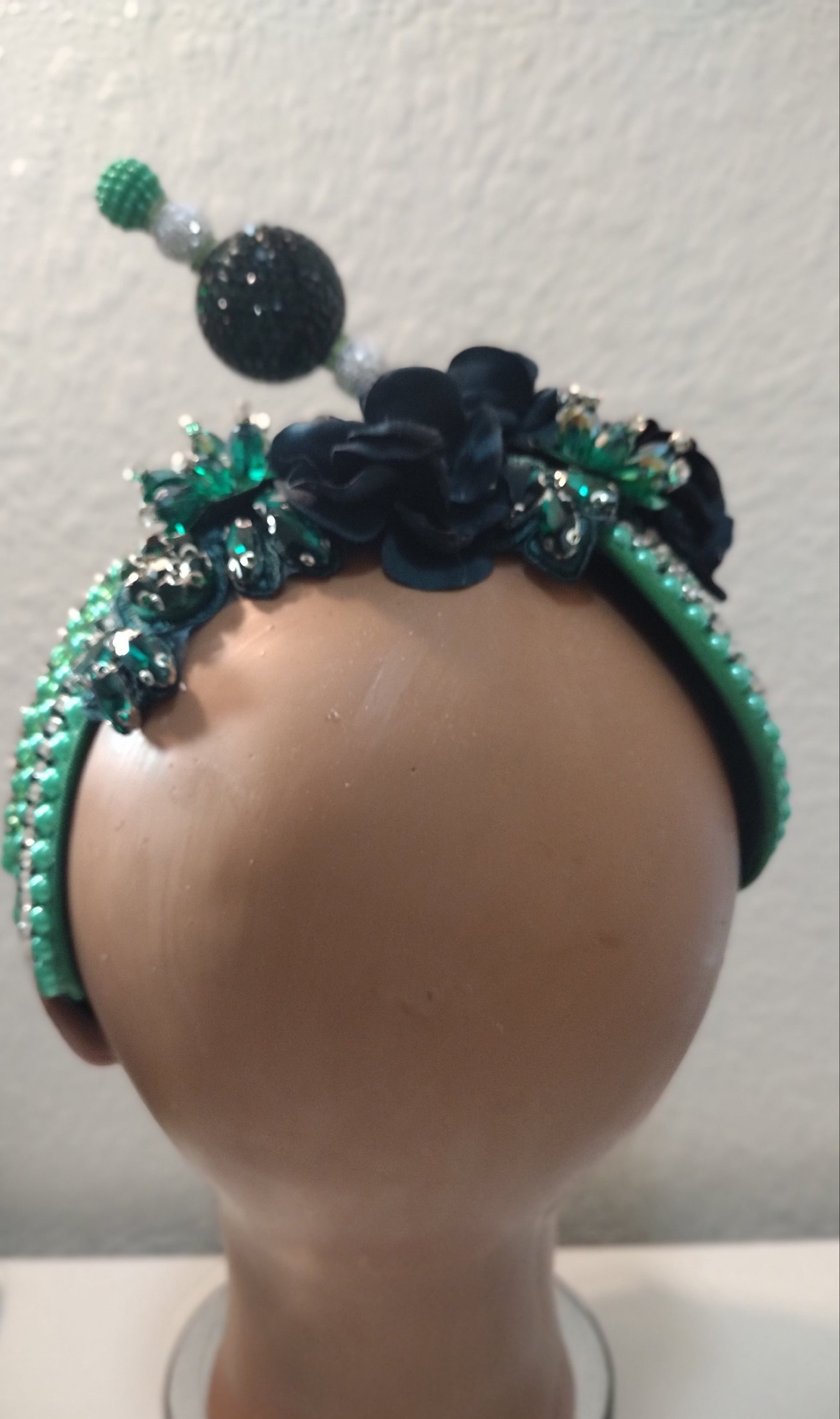 A decorative headband displayed on a mannequin head. The headband features dark navy blue fabric flowers, green beaded trim, and clear rhinestone embellishments.