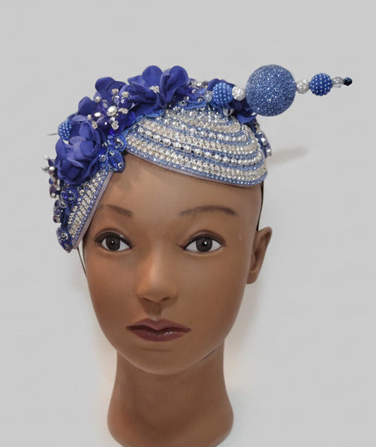 A blue and silver beaded headpiece with floral embellishments and a dangling beaded ornament, displayed on a mannequin head stand against a plain wall.