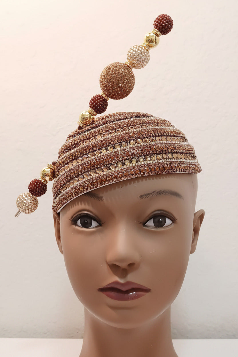 A close-up of a mannequin head wearing a heavily embellished brown and gold beaded cap with dangling rhinestone beads, photographed against a plain light wall.
