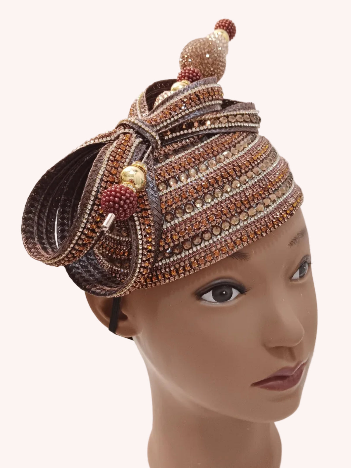 A highly embellished traditional African headwrap (dodo) displayed on a mannequin head. The wrap features intricate braided patterns in brown, beige, and gold, decorated with red and gold beaded tassels and a central beaded top ornament, set against a plain white wall background.