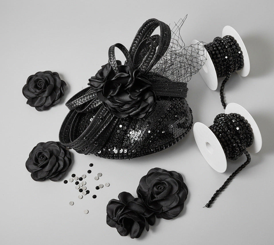 Black sequined fabric trim and rosette flowers displayed on a white surface against a textured beige wall, with a spool of beaded black ribbon standing behind the fabric pieces.
