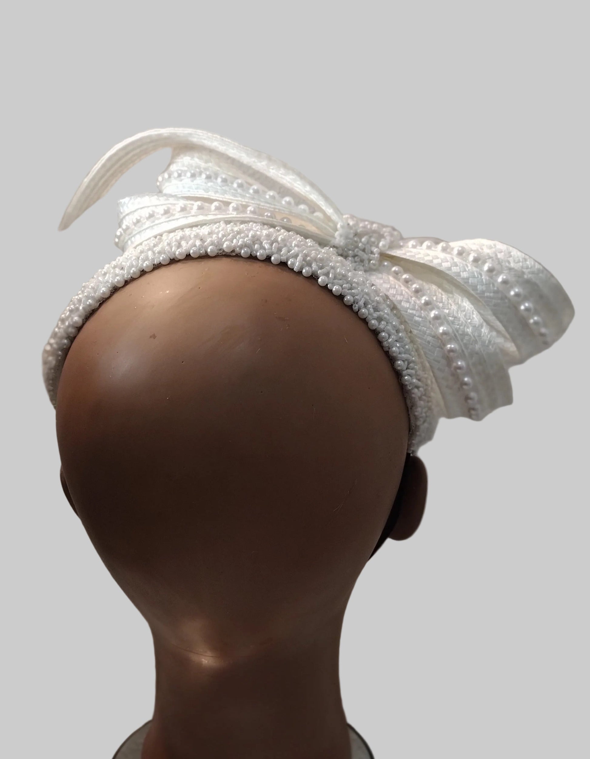 A white feathered fascinator hat with pearl trim displayed on a brown mannequin head against a solid gray background.