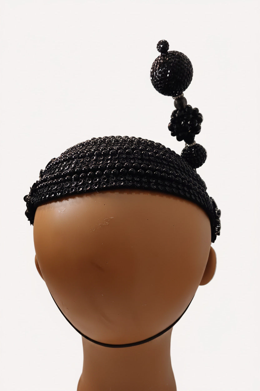 A black beaded headband with three vertical beaded tassels on the side, displayed on a mannequin head against a textured off-white wall.