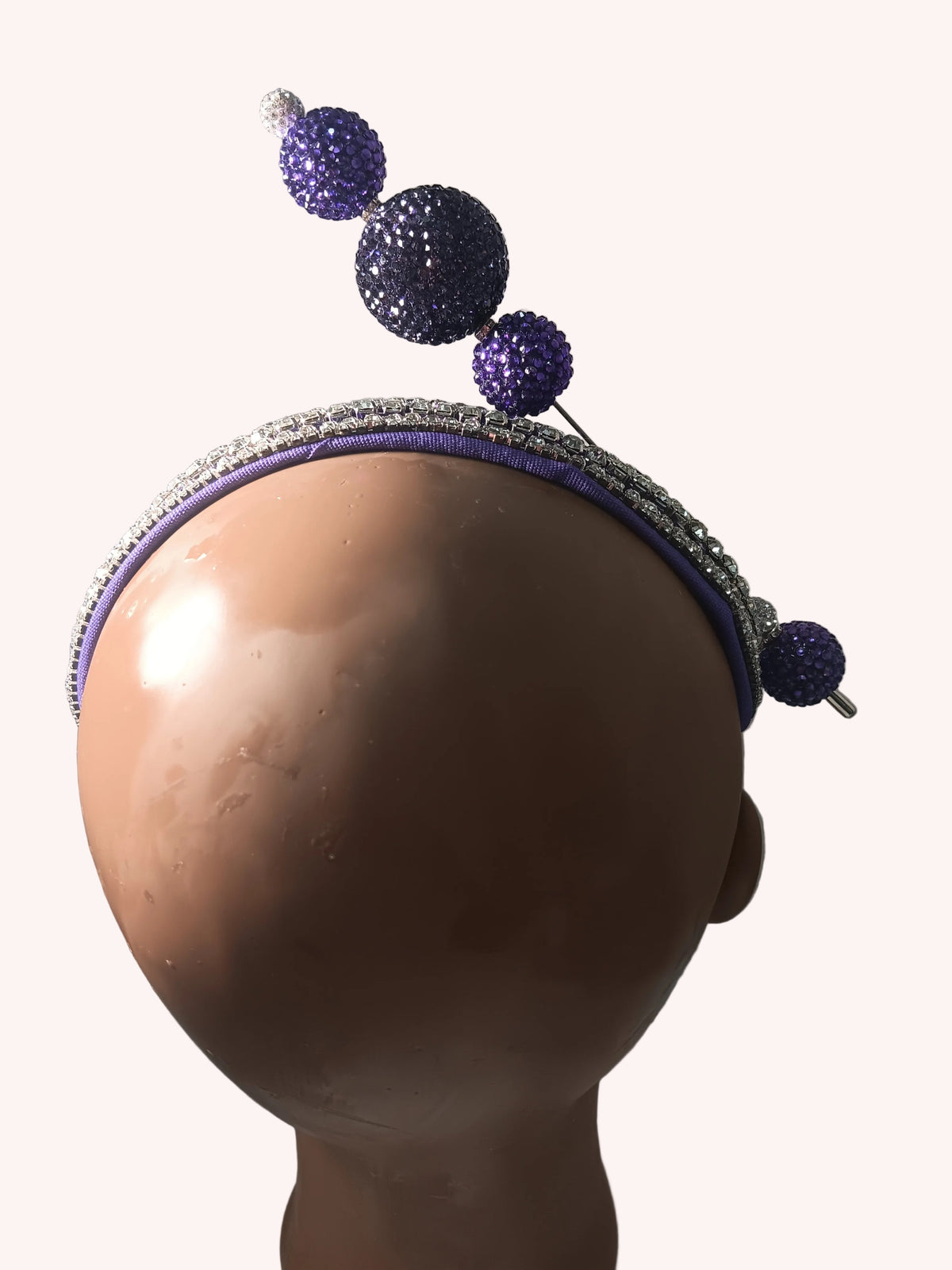 A purple and silver rhinestone headband with decorative purple rhinestone pom-poms, displayed on a mannequin head against a plain wall.