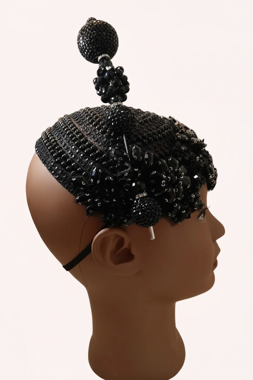A black beaded and sequined skullcap with a tall central bead tower and side embellishments, displayed on a mannequin head against a plain light wall.
