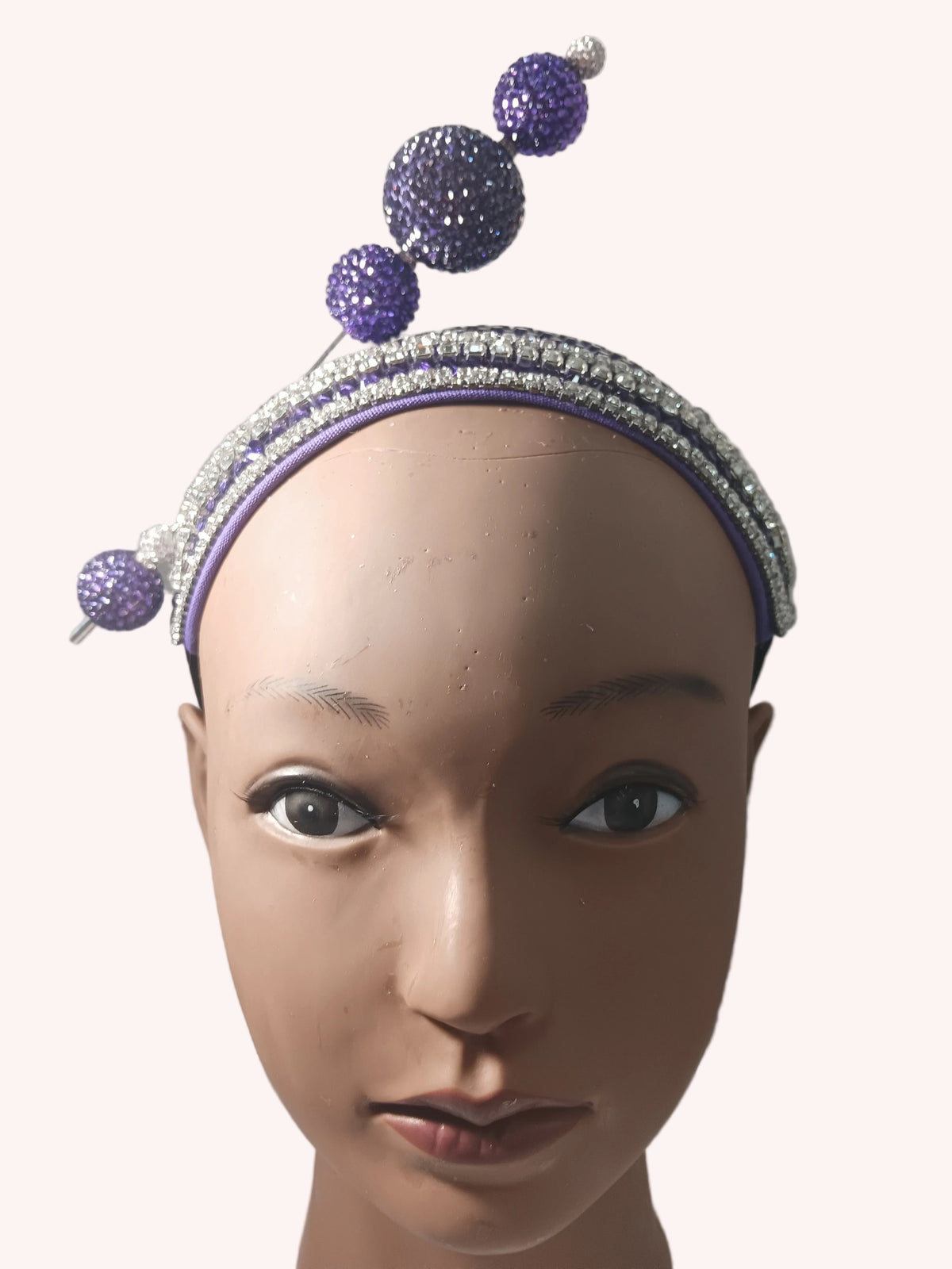 A purple and silver beaded headband displayed on a mannequin head. The headband features a wide silver rhinestone-embellished band, a purple inner trim, and three large purple rhinestone-covered spherical beads on top, with two matching purple beads on the sides.