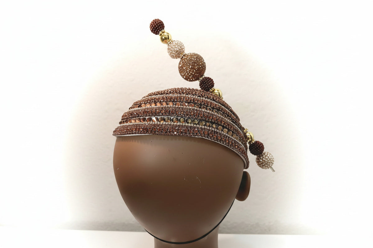 A beaded and rhinestone headpiece displayed on a brown mannequin head against a plain off-white wall. The headband features multiple horizontal rows of brown and clear rhinestones, with a diagonal top strand decorated with brown and clear beads, gold-toned spacer beads, and a dangling rhinestone tassel.