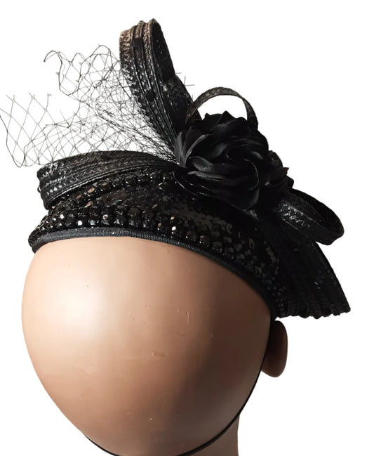 A black fascinator hat with a large fabric rose, sequined band, and attached netting, displayed on a beige mannequin head against a plain light wall.