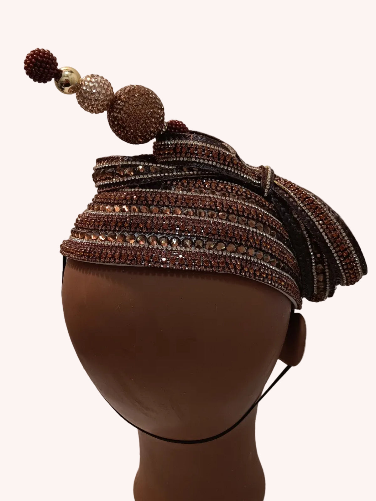 A brown beaded and sequined headpiece with decorative pom-poms and textured spherical accents, displayed on a brown mannequin head against a white wall.
