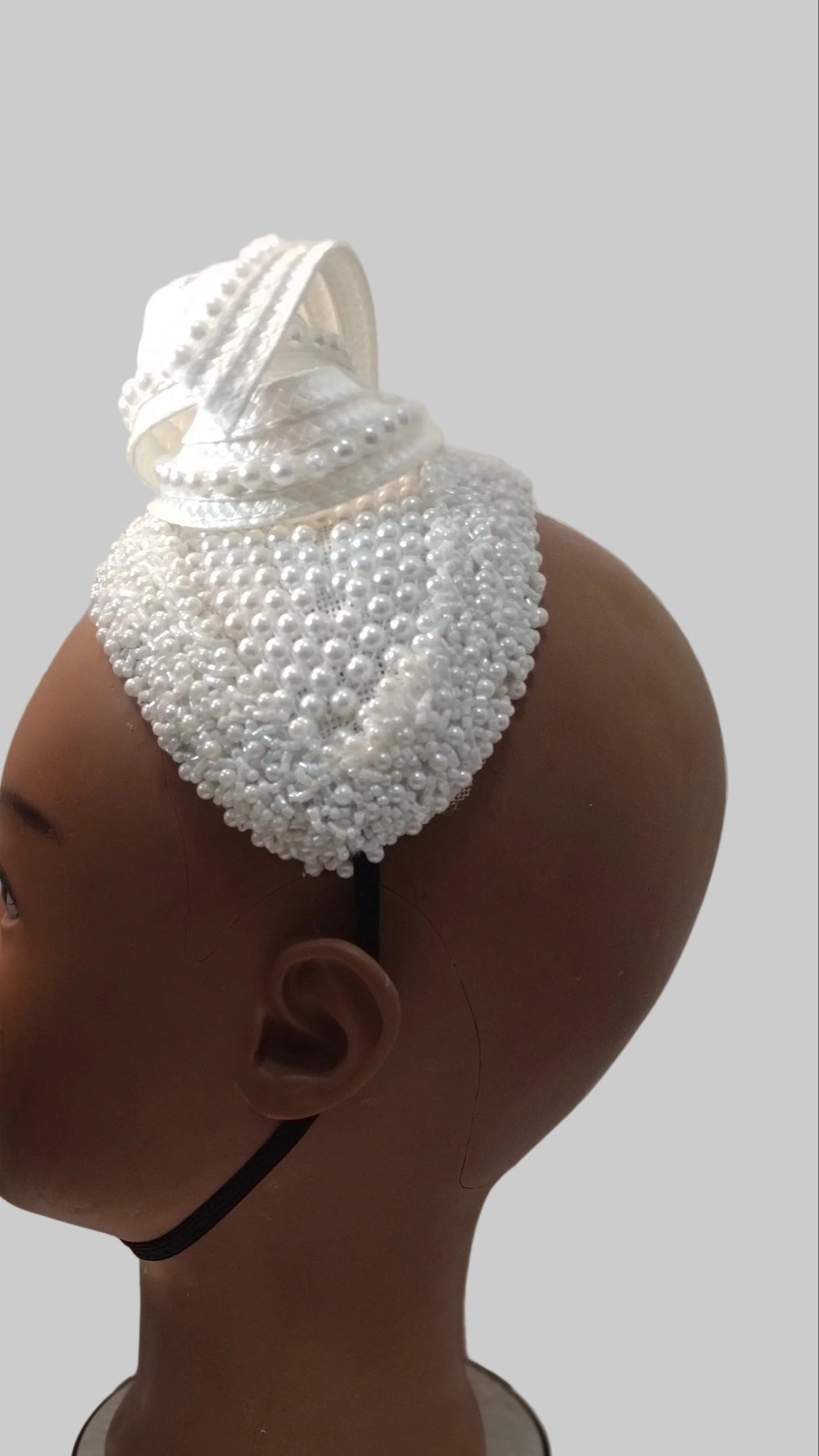 A white beaded and lace baby head covering cap displayed on a brown mannequin head, shown in profile against a textured off-white wall.